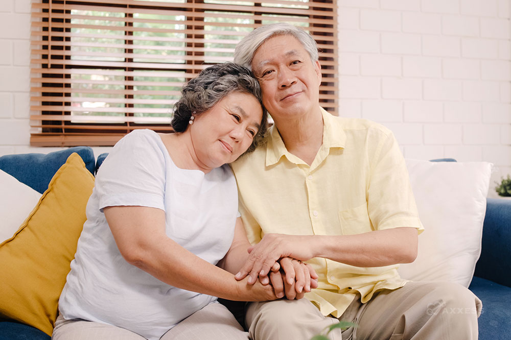 An older Asian couple is sitting on a couch. The woman is resting her head on the man's shoulder and they are holding hands.