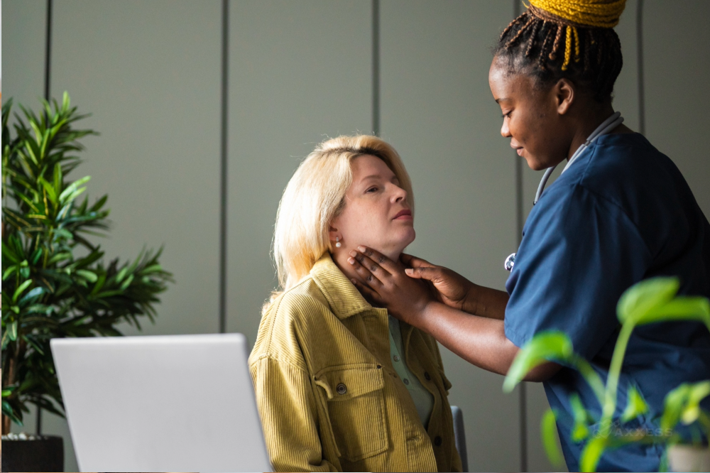 A female nurse in blue scrubs is checking a female patient's throat. The patient is sitting in a chair and wearing a yellow jacket. There is a laptop on the table next to them.