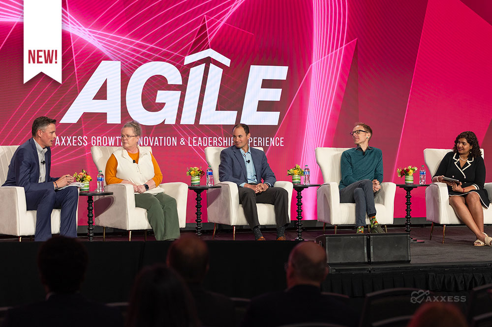 A panel discussion at a conference. There are five people on the panel, four men and one woman. They are all sitting in chairs on a stage. There is a large screen behind them that says "Agile."