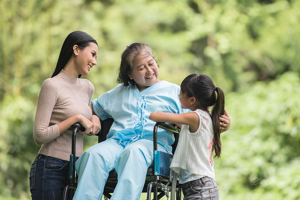 An elderly woman in a wheelchair outside looks fondly at her granddaughter, and her adult daughter looks on.