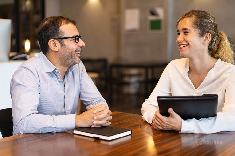 A male and female business professional sit at a table and smile at each other.