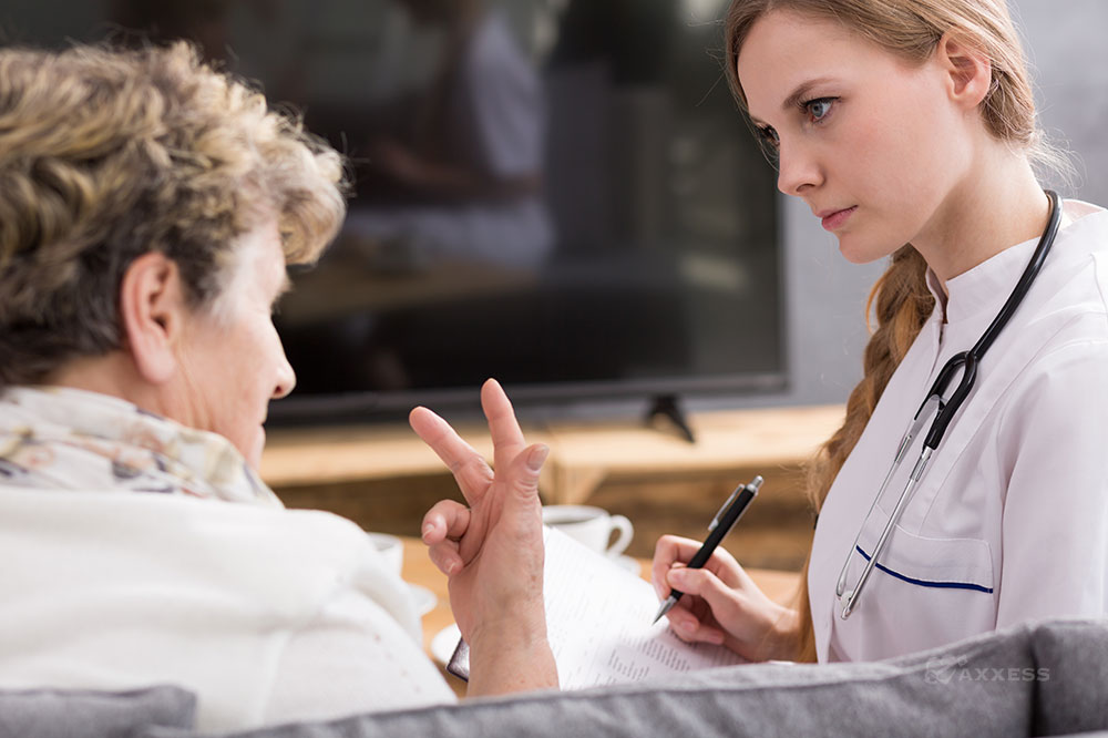 A blonde clinician holds a pen and looks at an older female patient who is speaking and gesturing with her hand.