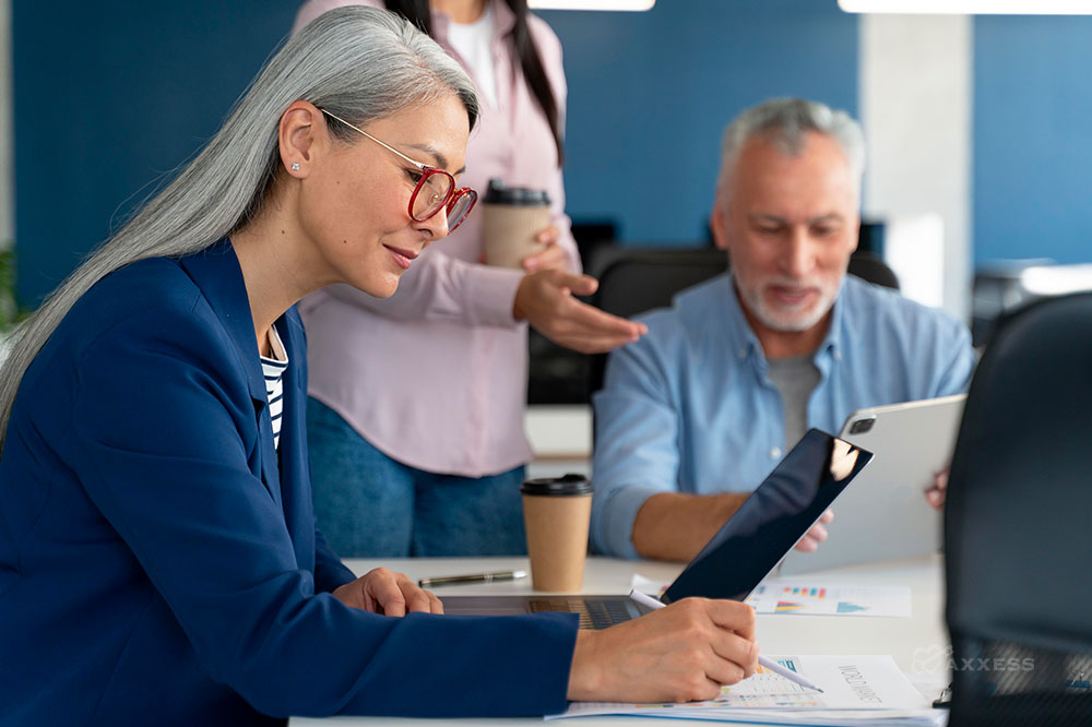 A woman with grey hair and glasses looks at a computer and graphs while sitting at a table