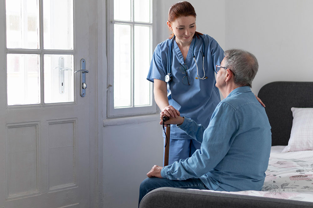 A female clinicians stands and assists a sitting male patient with his cane.