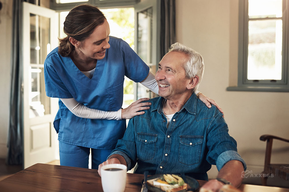 Female clinician in blue scrubs puts her hands on the shoulders of an elderly man sitting at a breakfast table