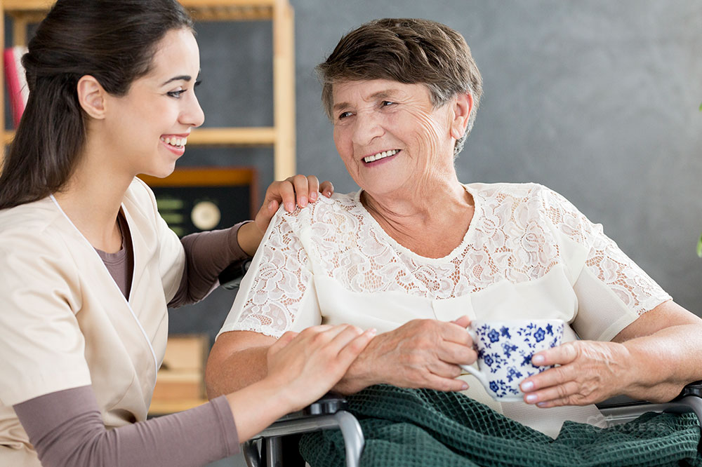 A female nurse sits next to an elderly female woman holding a teacup and smiles