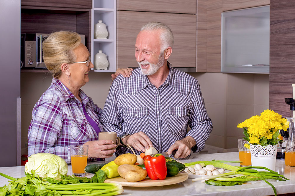 An elderly couple smiles at each other while standing at the kitchen counter preparing vegetables