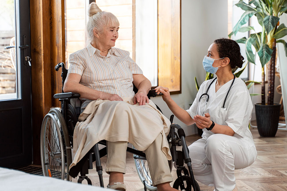 A home health clinician kneels next to an elderly patient in a wheelchair