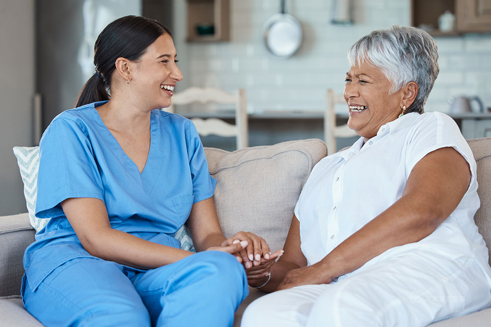 A female clinician in blue scrubs sits on a couch with an elderly client smiling