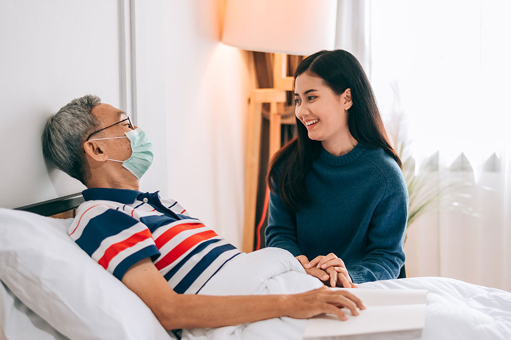A young woman smiles while sitting next to bed where an older man wearing a mask is laying.