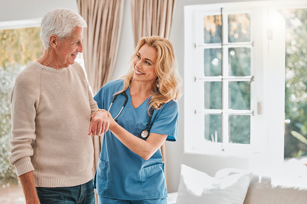 A female clinician in blue scrubs stands and holds the arm of an elderly man
