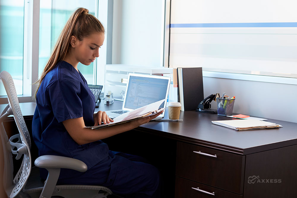 A home care administrator in blue scrubs sits at a desk and looks over paperwork