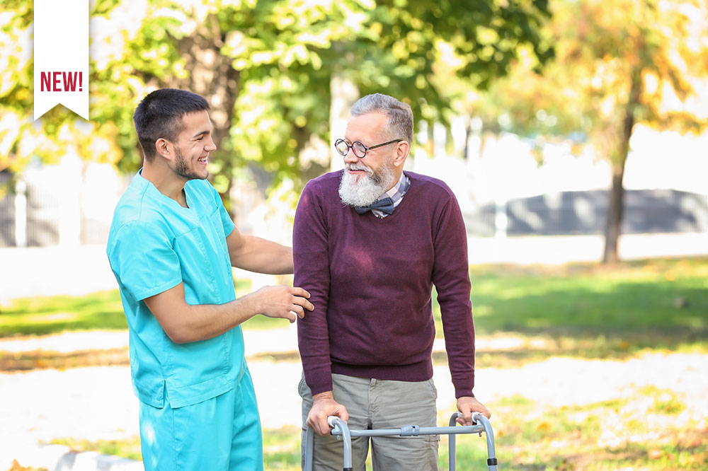 A male clinician in teal scrubs assists an elderly man in a maroon sweater walking outside with a walker
