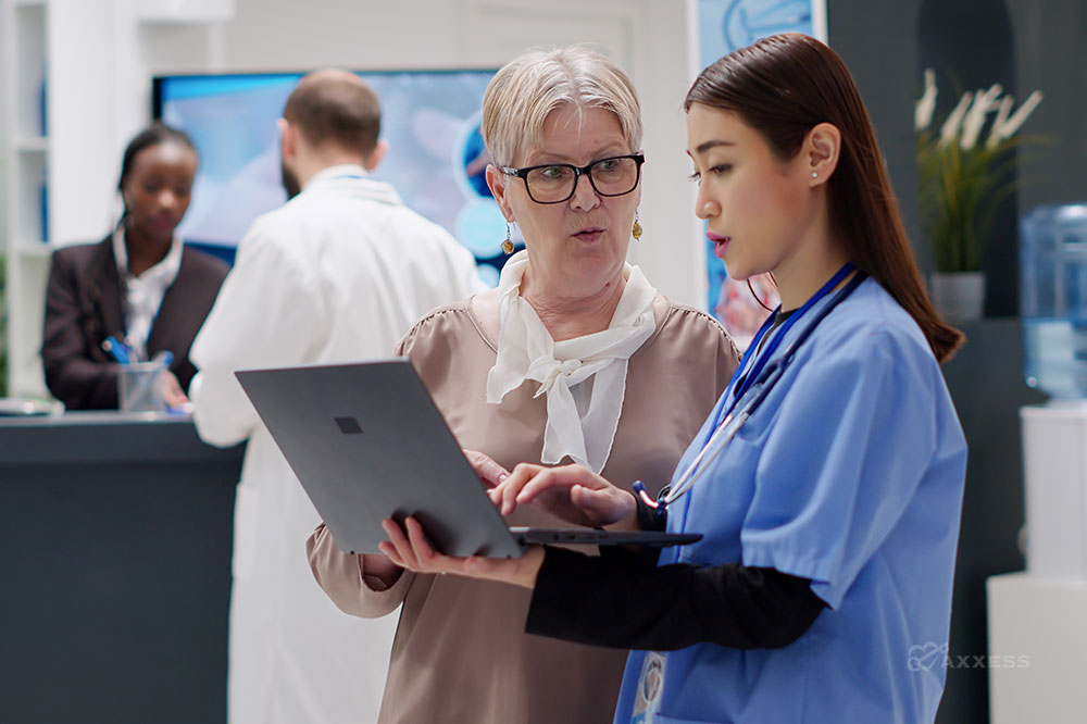 A nurse holding a laptop while another woman, who is the office manager, explains how to use the new technology for patient care. In the background a doctor speaks to a receptionist.