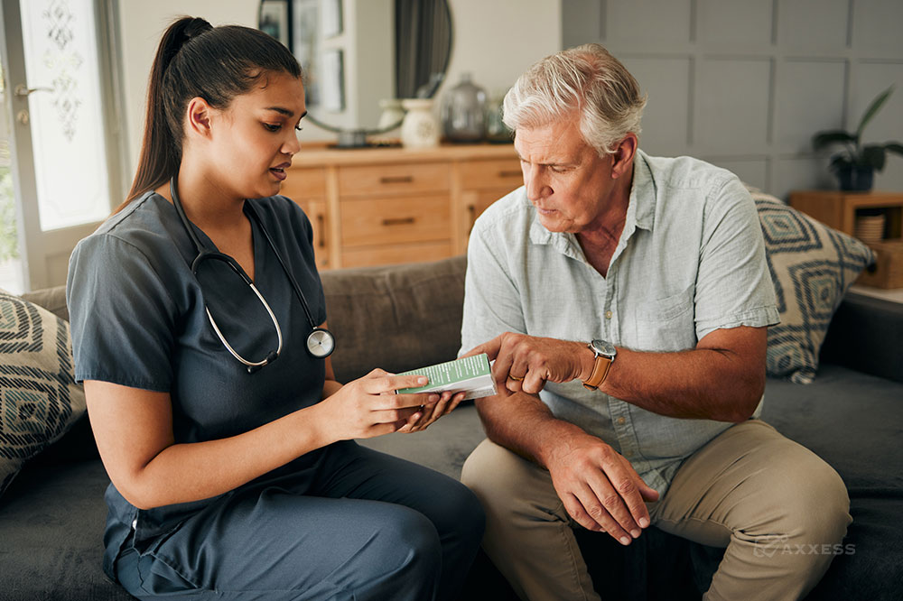 A female nurse is sitting on a couch next to a senior male patient. She is holding a pill bottle and explaining the prescription to him.