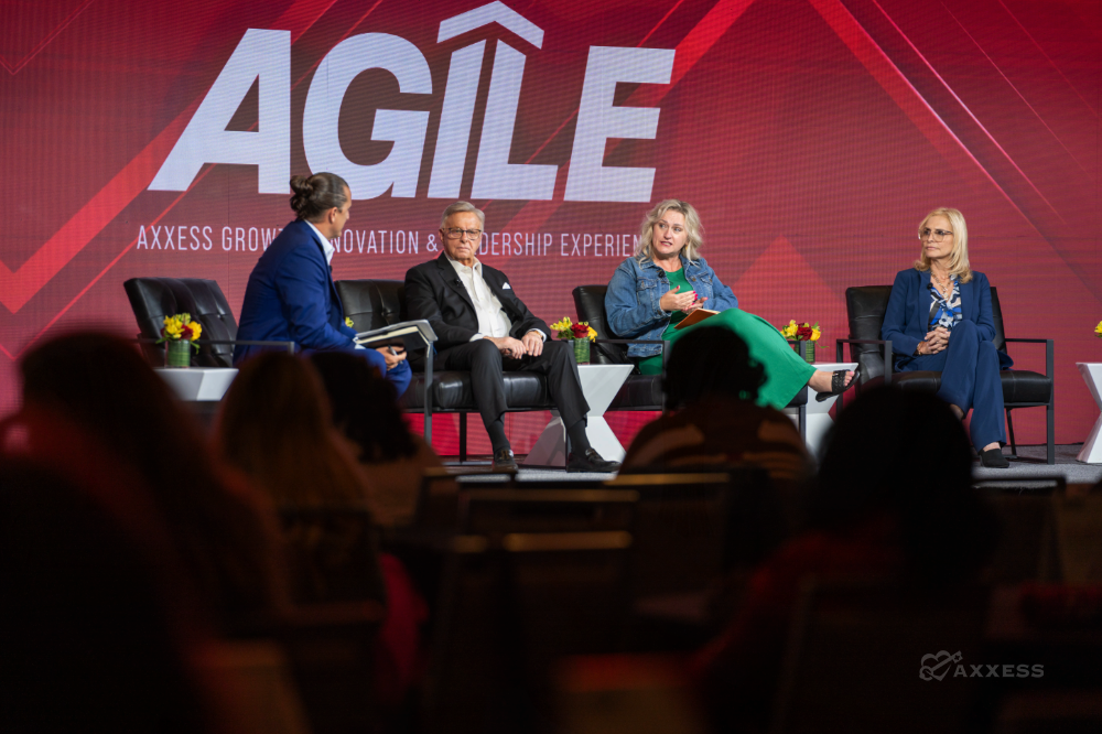 A panel of four speakers is seated on a stage in front of a large red and white sign. The speakers are arranged in a row of black chairs, with a man on the left, a man in the center-left, a woman in the center right, and a woman on the right. The audience is visible in the foreground, mostly blurred, with people seated in rows of chairs. The lighting is bright on the stage, and the overall tone of the image is professional and informative. The sign behind them reads "Axxess Growth Innovation & Leadership Experience".
