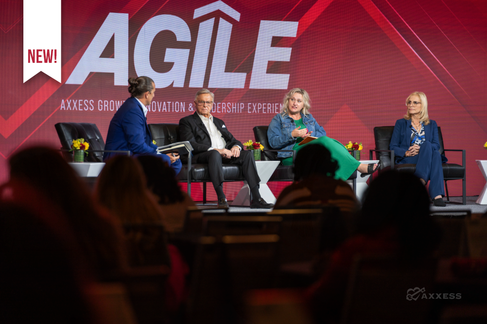 A panel of four speakers is seated on a stage in front of a large red and white sign. The speakers are arranged in a row of black chairs, with a man on the left, a man in the center-left, a woman in the center right, and a woman on the right. The audience is visible in the foreground, mostly blurred, with people seated in rows of chairs. The lighting is bright on the stage, and the overall tone of the image is professional and informative. The sign behind them reads "Axxess Growth Innovation & Leadership Experience".