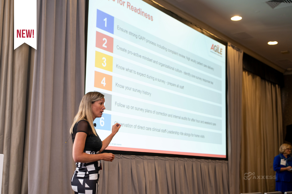 A woman presents in a conference room, standing beside a projection screen displaying a slide titled Tips for Readiness. The slide lists six color-coded items related to survey readiness, including operational processes, preparation calendars, survey history, and staff alignment. A laptop sits on a podium, and another person stands in the background. Rows of chairs face the presenter.