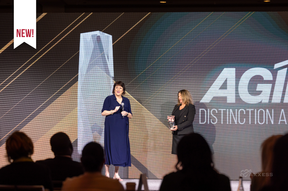 Award ceremony at the 2025 Axxess AGILE conference in Dallas, Texas, featuring the presentation of the AGILE Distinction Award on stage. Two individuals stand at the center, one speaking into a microphone, the other holding the trophy, while a digital display and audience are visible in the background.