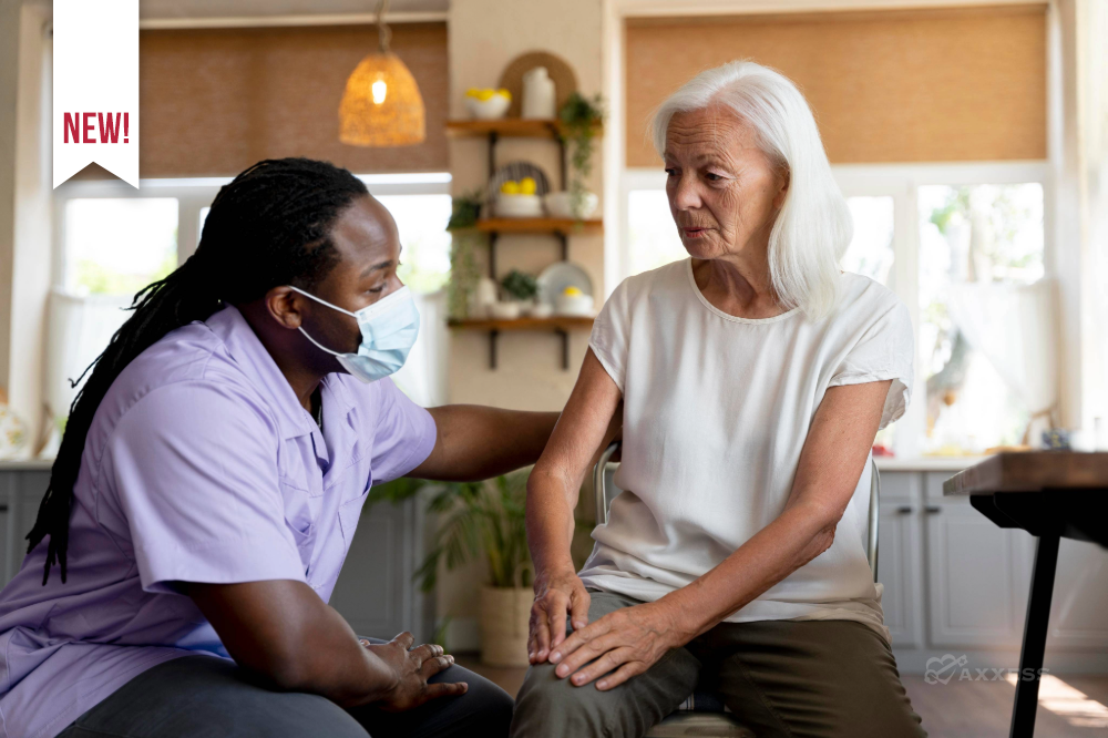 A healthcare professional in scrubs and a face mask provides supportive, compassionate care to an older adult in a bright home setting, gently holding their arm during a conversation. The scene highlights home‑based care, senior support and caregiver&ndash;patient connection.