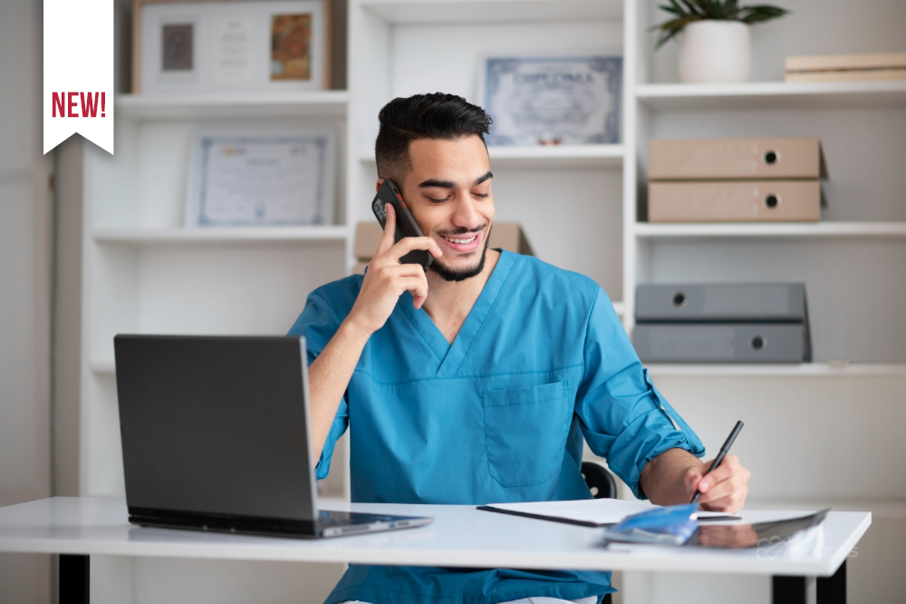 A healthcare professional wearing blue scrubs sits at a desk, talking on a smartphone while writing in a notebook. An open laptop, tablet, and papers are spread out on the desk, and shelves with binders, certificates, and decor are visible in the background.