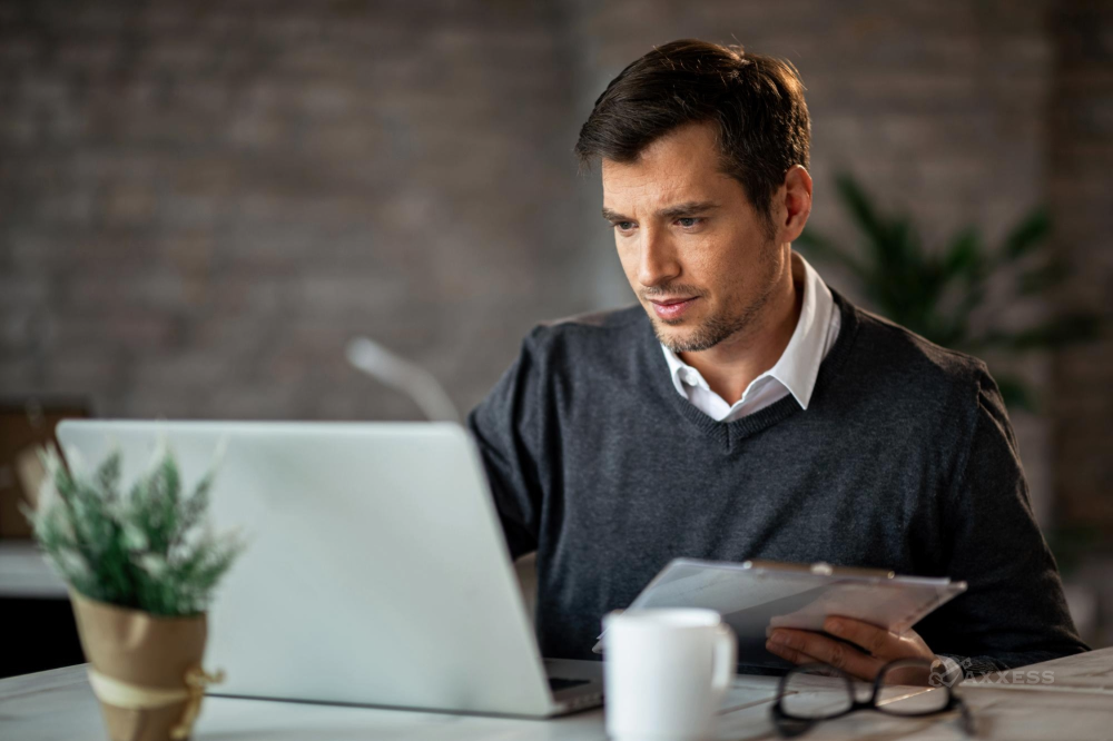 A person sits at a desk working on a laptop while holding a clipboard. A coffee mug, eyeglasses, and a small potted plant are on the desk, creating a focused office workspace.
