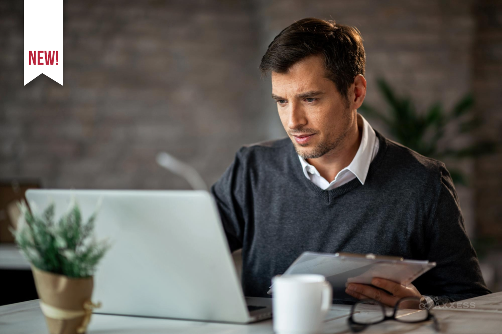 A person sits at a desk working on a laptop while holding a clipboard. A coffee mug, eyeglasses, and a small potted plant are on the desk, creating a focused office workspace.