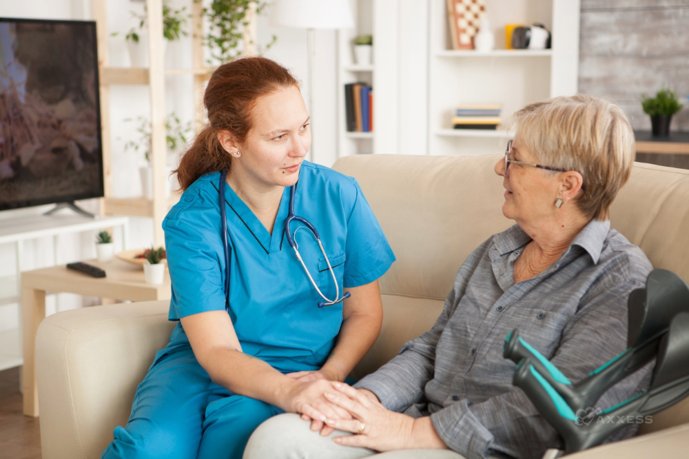 A healthcare professional in blue scrubs sits on a couch beside an older adult, gently holding their hand and speaking with them in a warm, supportive manner. They are in a cozy living room with a television, bookshelves, and a potted plant in the background, conveying a calm, home‑based care setting.