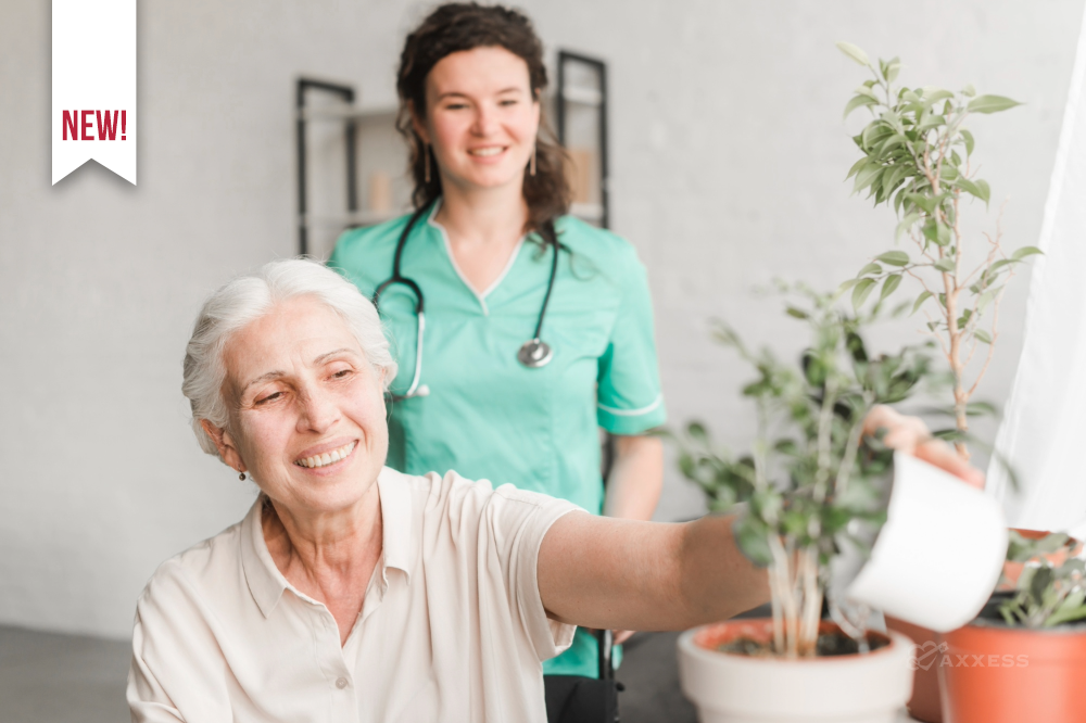 An older adult sits indoors watering a potted plant with a white watering can while a healthcare professional in green scrubs stands behind them, offering support. Several plants surround them in a bright, comfortable room with a window and light-colored walls.